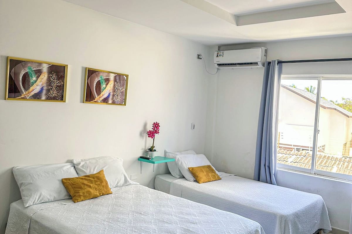 Habitación de hotel con dos camas, una lámpara en el techo, un equipo de aire acondicionado, una ventana con cortinas y dos cuadros colgados en la pared.