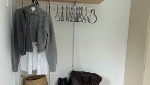Pieni huone, jossa on sänky, puinen hylly, jossa on siististi taiteltuja vaatteita, henkariteline takkeja varten, punottu kori, käsilaukku, kangaskassi ja pari lenkkareita.
