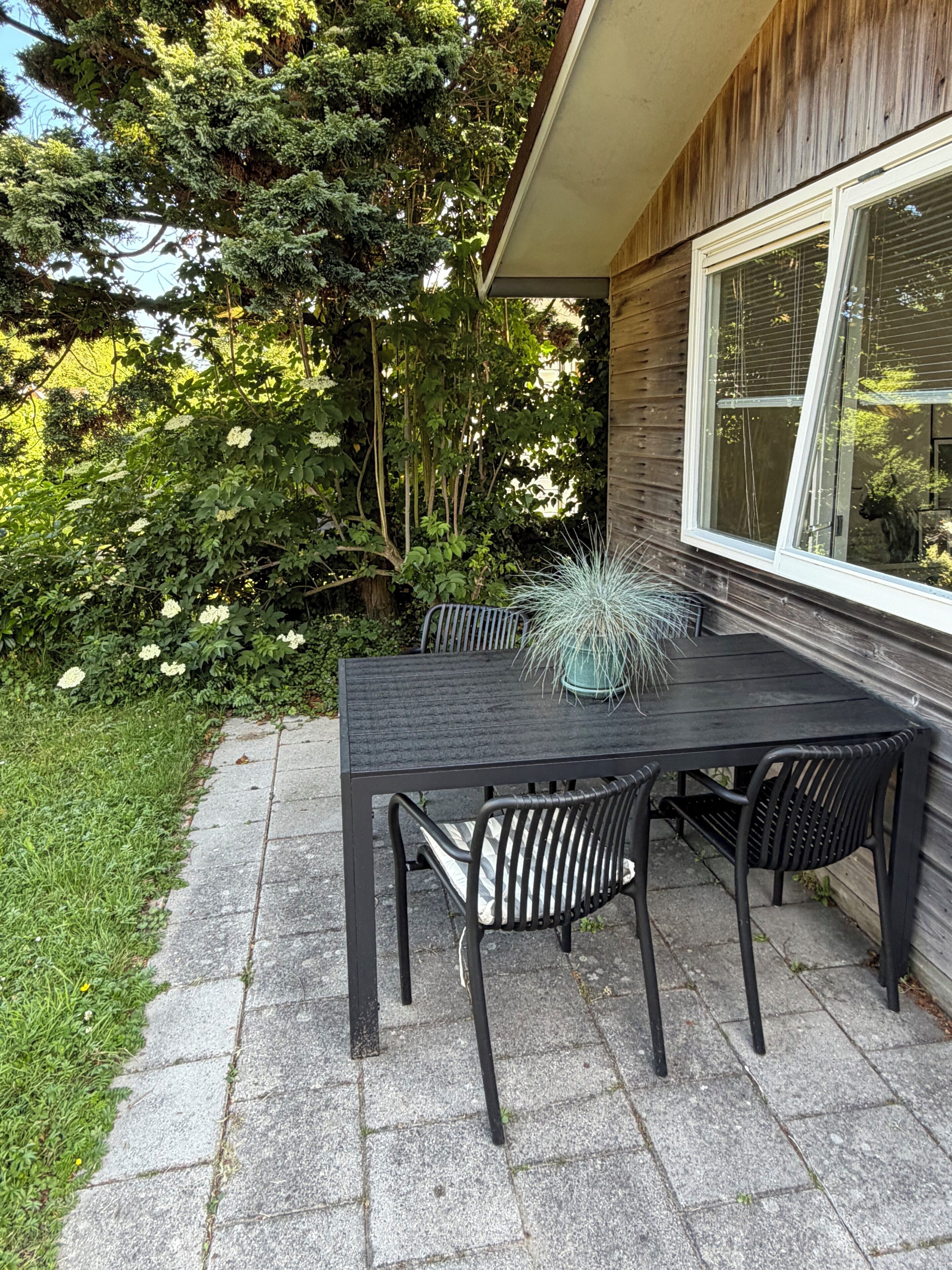 Une terrasse extérieure avec une table et des chaises noires, entourée d’une végétation luxuriante et d’un bâtiment en bois.