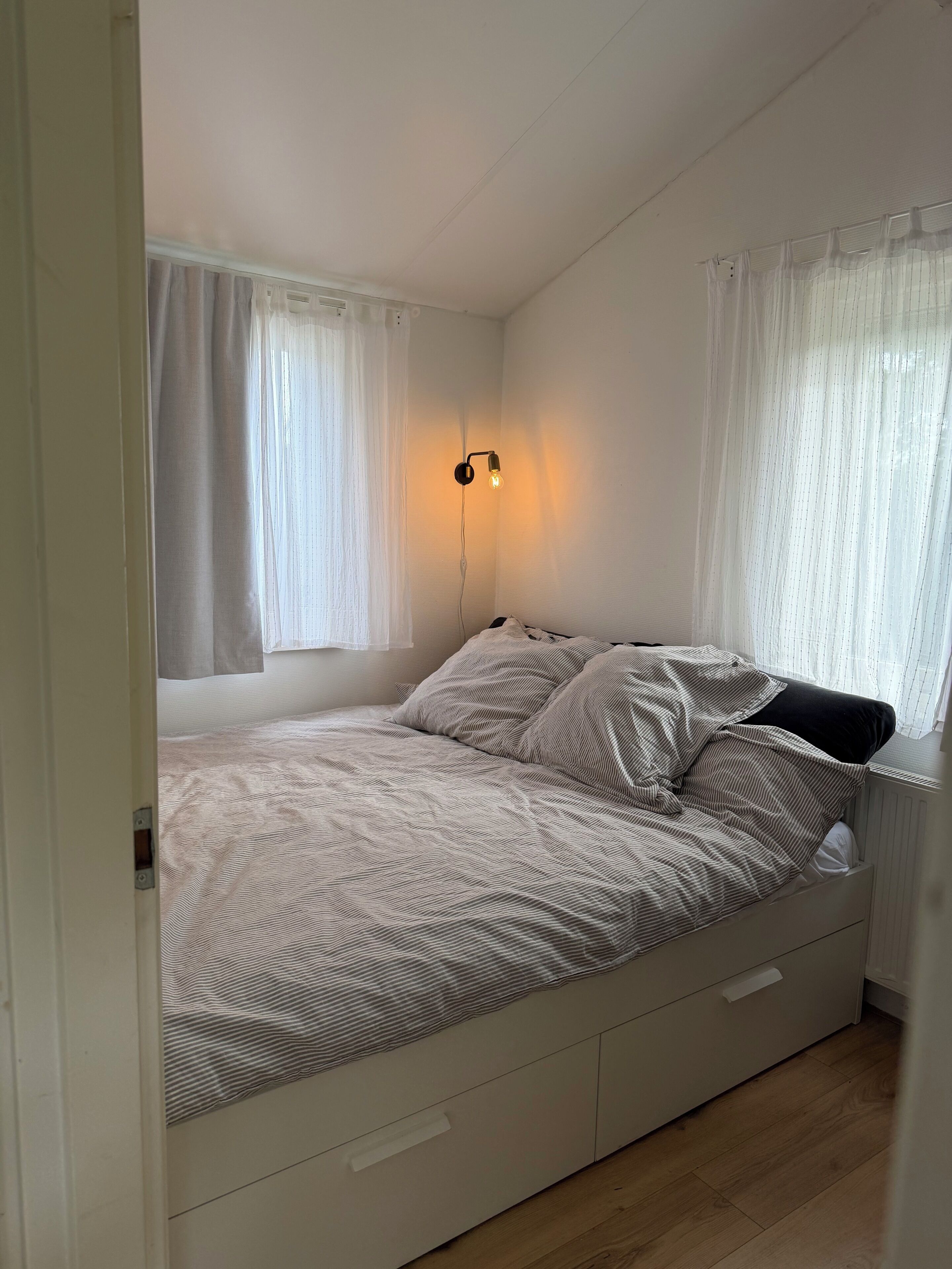 A bedroom with a bed, a nightstand, a wall-mounted lamp, and a window with sheer curtains.