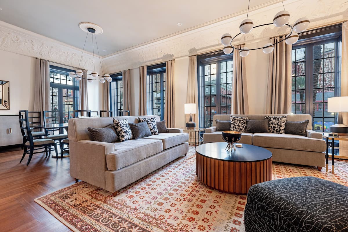 A spacious living room with two sofas, a coffee table, and a dining area with chairs and a table. Large windows provide views of the city.