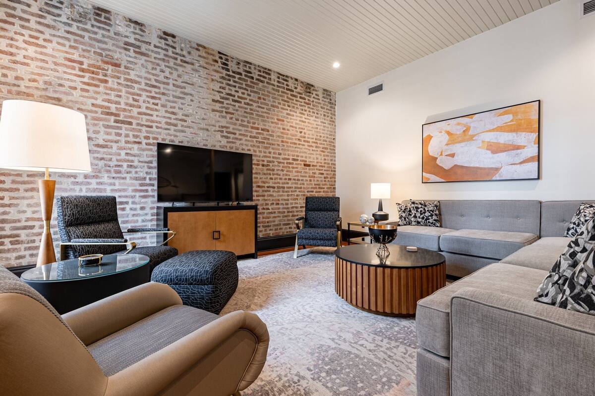 A modern living room with a brick wall, a flat-screen TV, a grey sofa, a patterned armchair, and a coffee table.