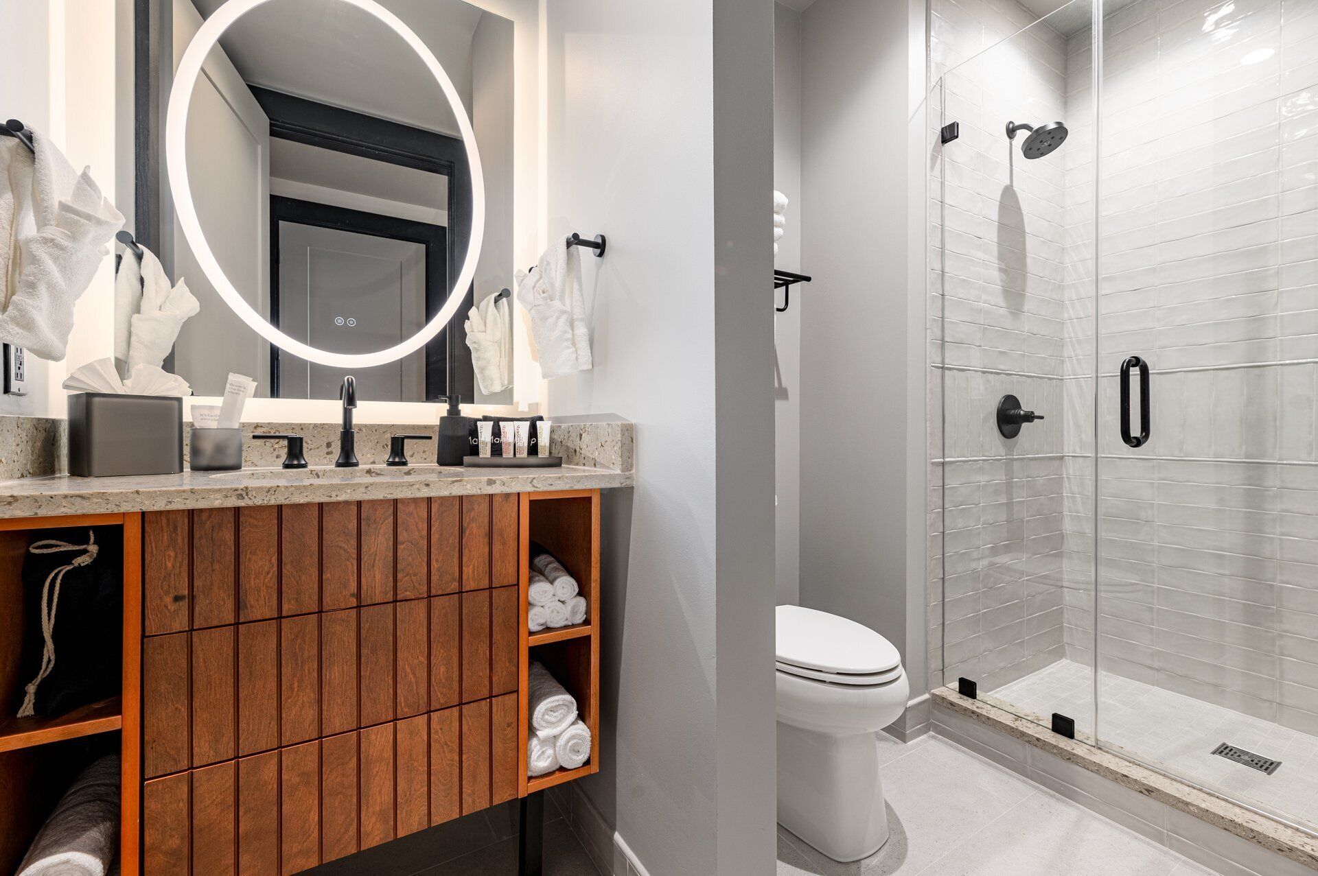 Een moderne badkamer met een houten wastafelmeubel, een ronde spiegel, een douche met glazen deur en een toilet.