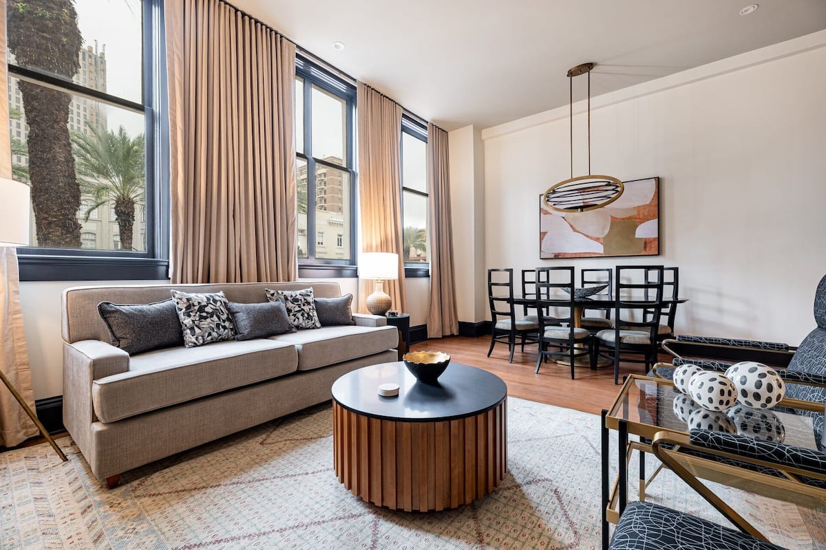 A modern living room with a sofa, a round coffee table, a dining area with chairs, and a large window with curtains.