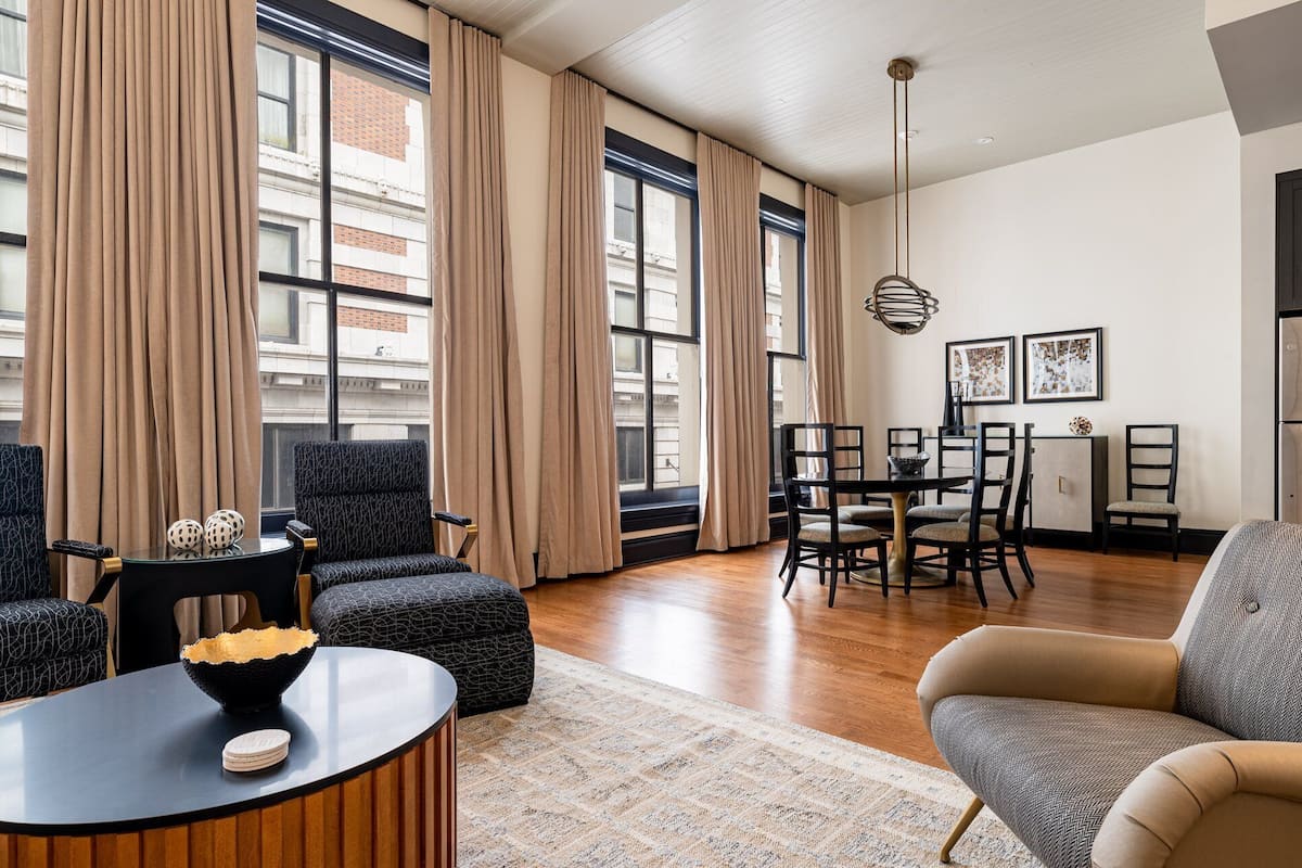 A spacious living room with a large window, a dining area with a table and chairs, and a seating area with a sofa and ottoman.