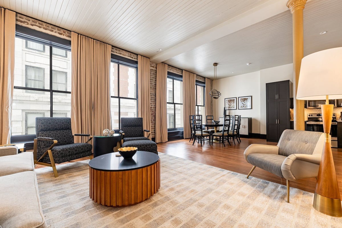 A spacious living room with a round wooden coffee table, two armchairs, a sofa, and a dining area with chairs and a table.