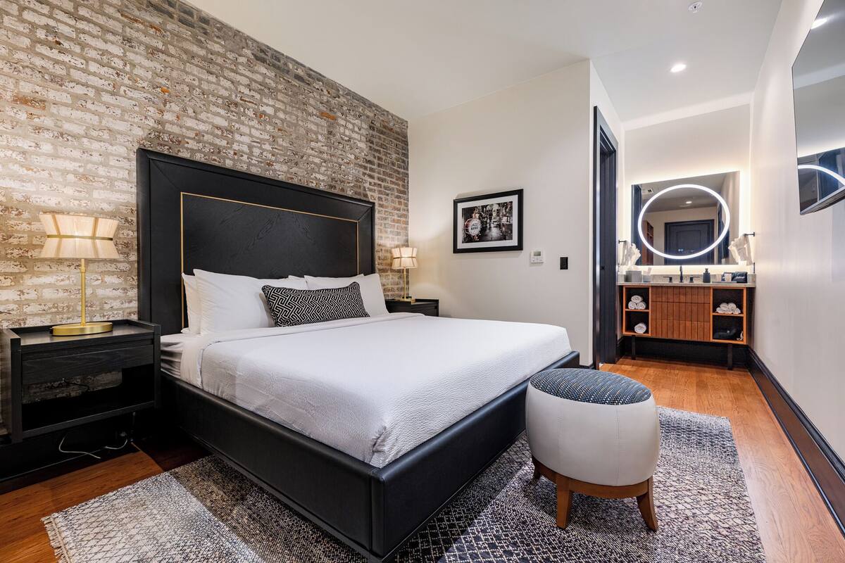 A modern bedroom with a brick wall, a large bed, a bedside table with lamps, a small stool, and a bathroom with a mirror and shelves.