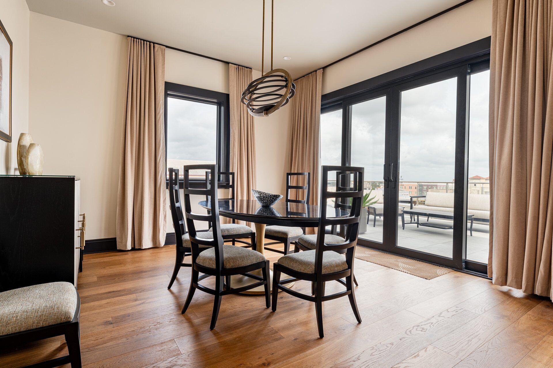 A modern dining room with a round table, six chairs, and a hanging light fixture. Large windows with curtains offer a view of the cityscape.