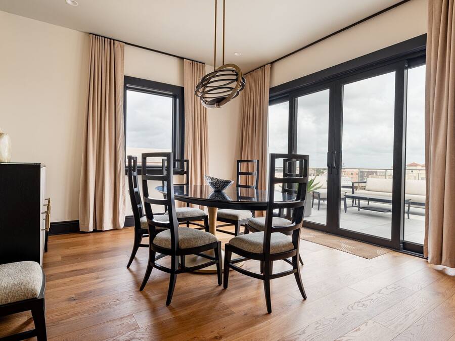 A modern dining room with a round table, six chairs, and a hanging light fixture. Large windows with curtains offer a view of the cityscape.