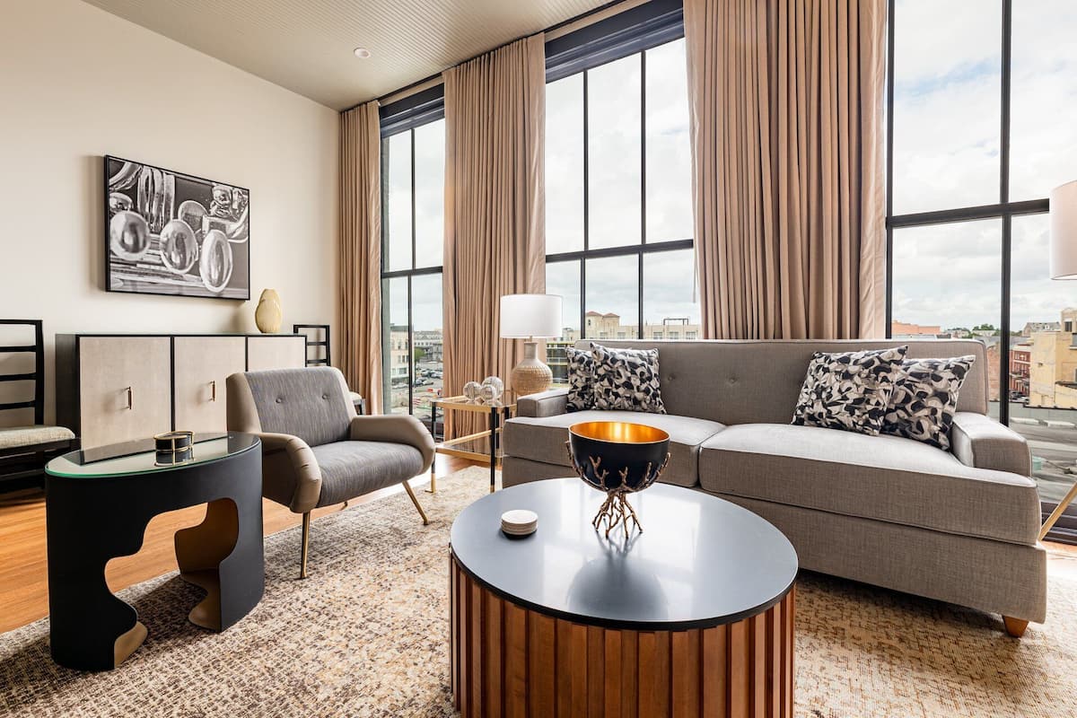 A modern living room with a grey sofa, a round coffee table, and a black chair. There is a large window with curtains, a painting on the wall, and a lamp on a side table.