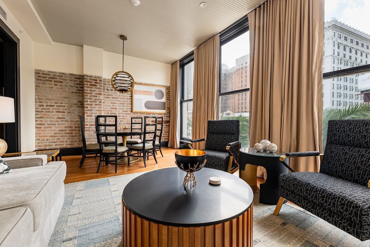 A modern living room with a brick wall, a round wooden coffee table, a black sofa, and a dining area with a table and chairs.