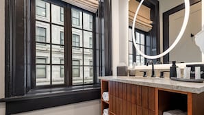 A bathroom with a large window, wooden vanity, and a mirror with a light fixture. - The Mayfair (New Orleans)