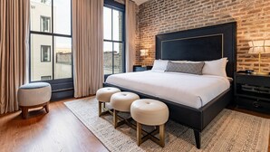A modern bedroom with a brick wall, large windows, a black headboard, and a wooden floor. - The Mayfair (New Orleans)