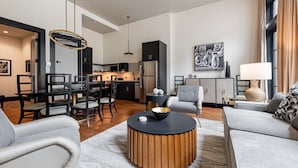A modern living room with a round coffee table, grey sofas, and a dining area with a black and gold chandelier. - The Mayfair (New Orleans)