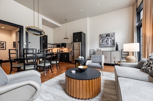 A modern living room with a round coffee table, grey sofas, and a dining area with a black and gold chandelier.