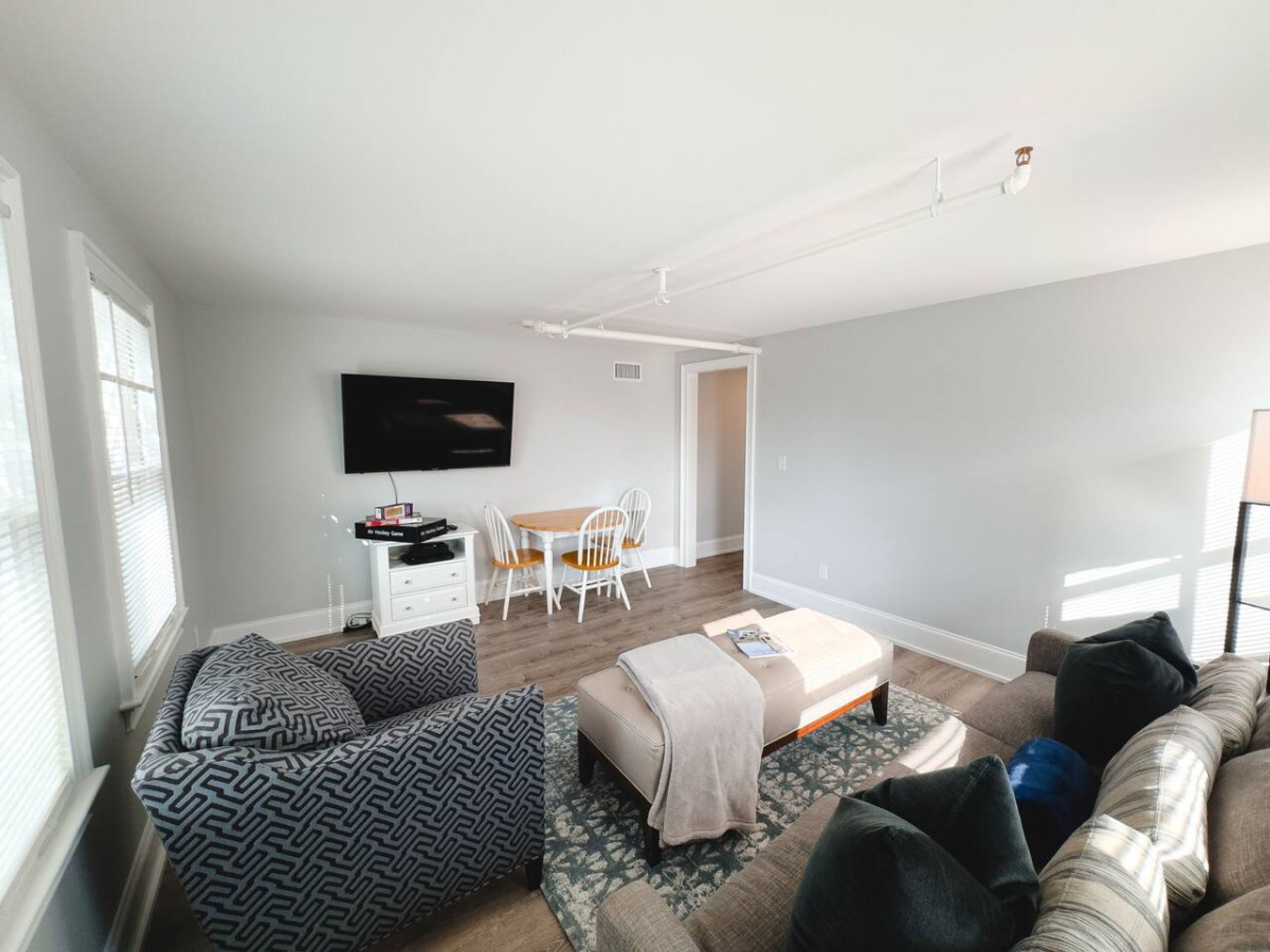 A modern living room with a sofa, a coffee table, and a TV mounted on the wall. There is a dining area with a table and chairs.