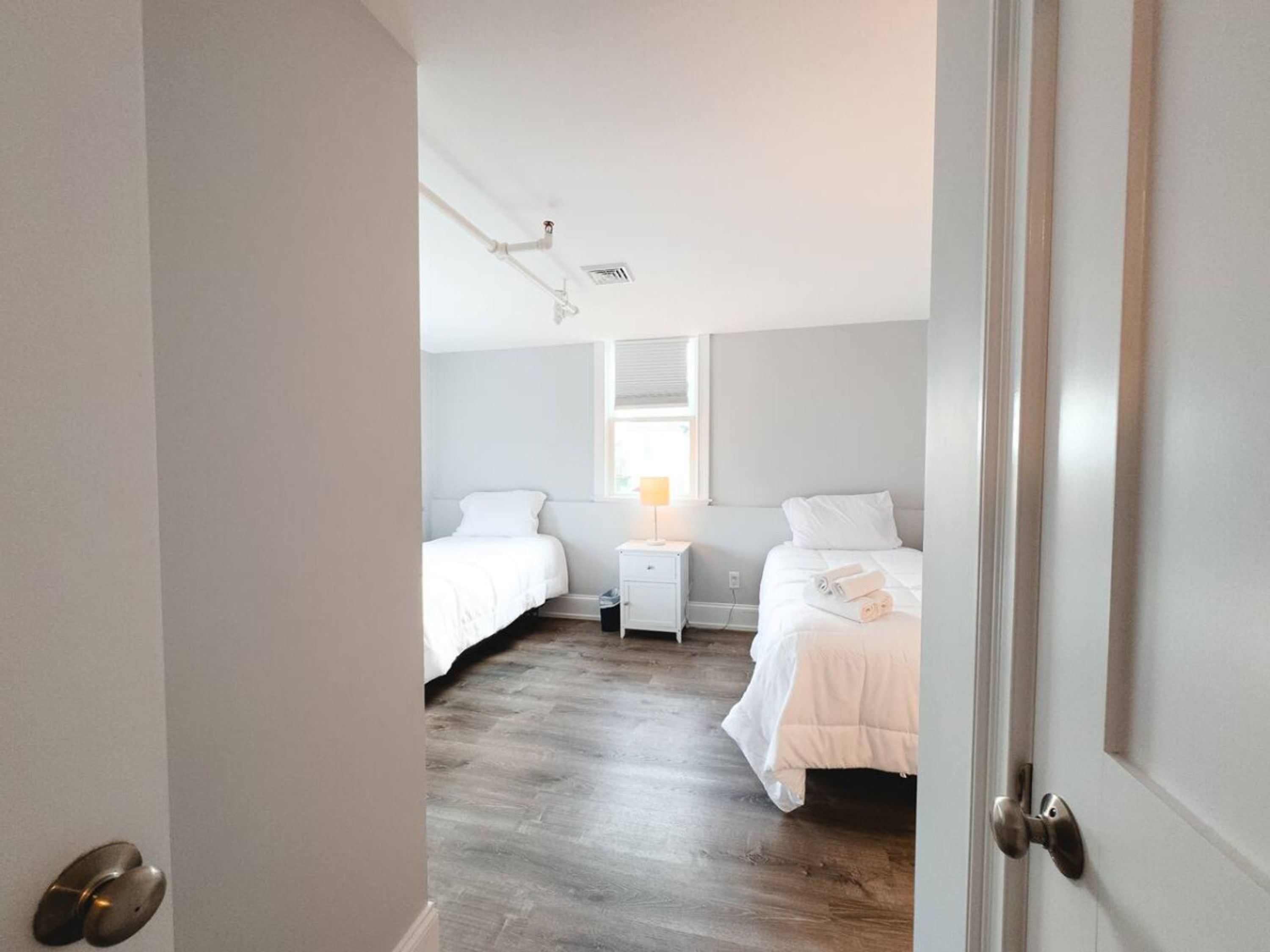 Une chambre avec deux lits, une fenêtre avec des stores, une table de chevet avec une lampe et une porte.