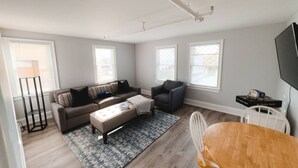 Un salon spacieux comprenant un canapé, un fauteuil et un pouf, une table à manger avec des chaises et un téléviseur.