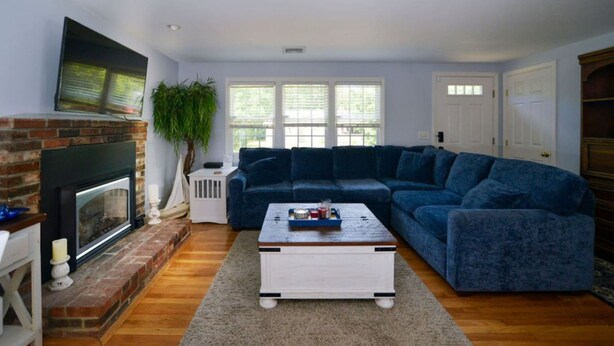 A living room with a brick fireplace, a blue sofa, a coffee table, and a large window with blinds. - Coastal Haven in West Dennis -15 (West Dennis)