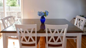 A dining area with a glass-top table, white chairs, and a blue vase with flowers. - Coastal Haven in West Dennis -15 (West Dennis)