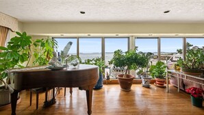 A spacious room with a grand piano, large windows offering a view of the sea, and various potted plants. - Coastal Charm Lobster Ch Teau (Swampscott)