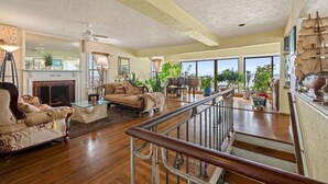 A spacious living room with a fireplace, a grand piano, and large windows offering a view of the outdoors. - Coastal Charm Lobster Ch Teau (Swampscott)