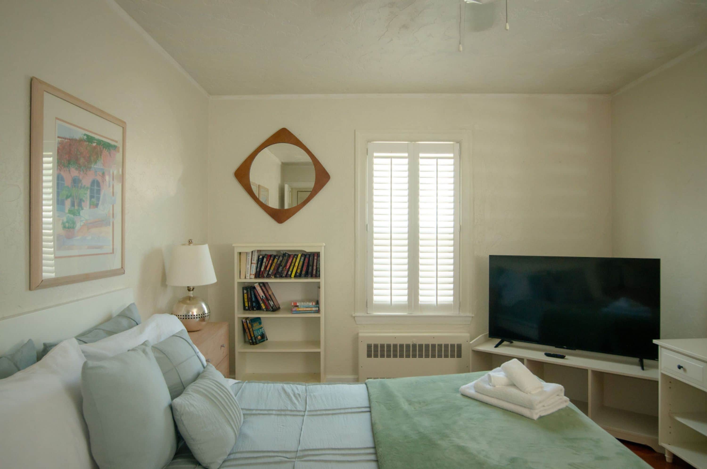 A bedroom with a bed, a TV, a bookshelf, a mirror, and a window with blinds.