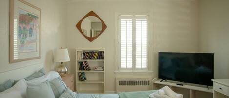 A bedroom with a bed, a TV, a bookshelf, a mirror, and a window with blinds.