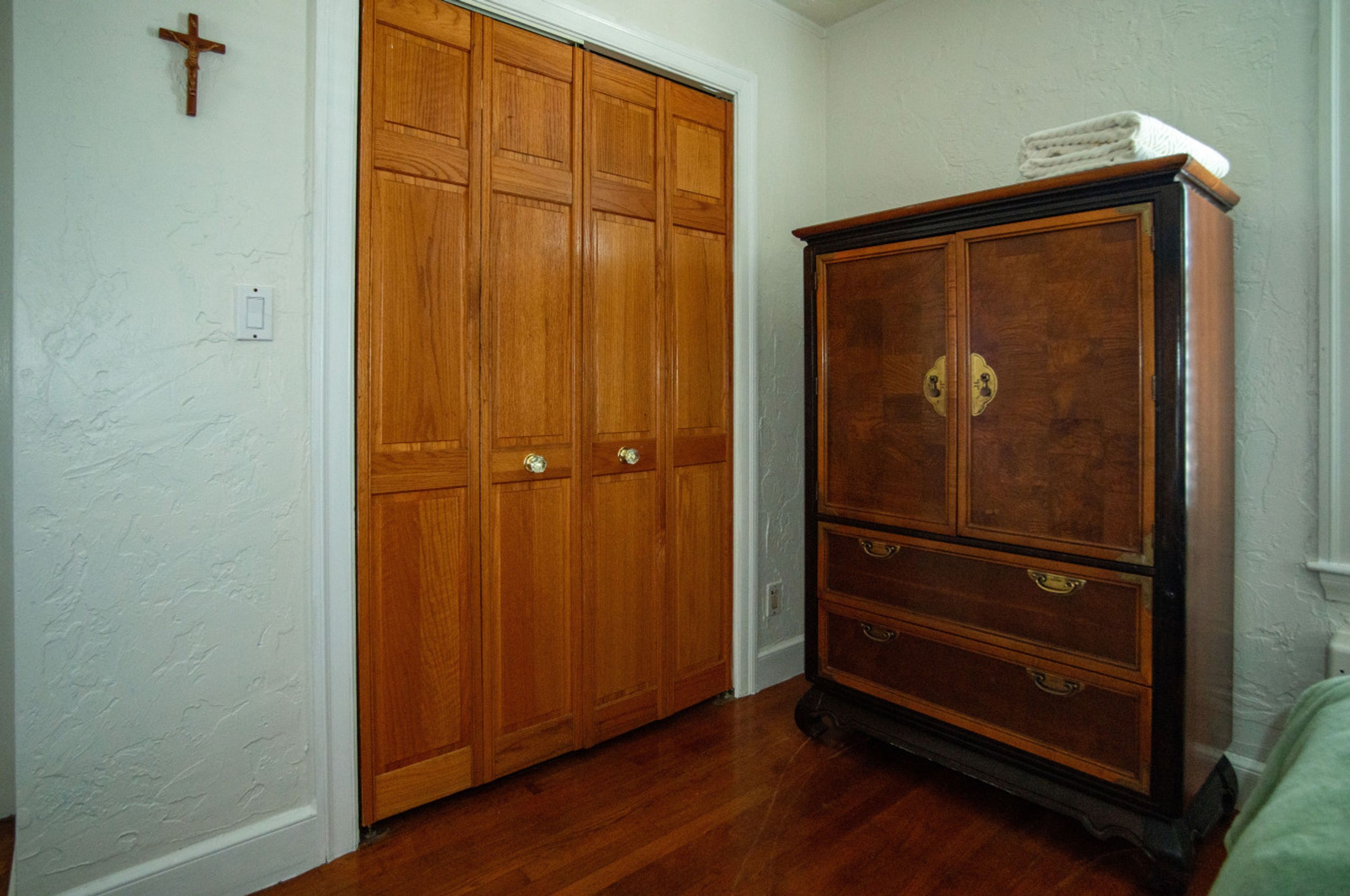 A room with a wooden wardrobe, a wooden cabinet, and a cross on the wall.