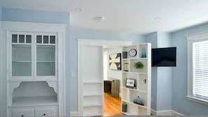 A hallway with a white door, a white shelving unit with decorative items, and a flat-screen TV mounted on the wall. - Enchanting Hidden Door Escape (Stoneham)