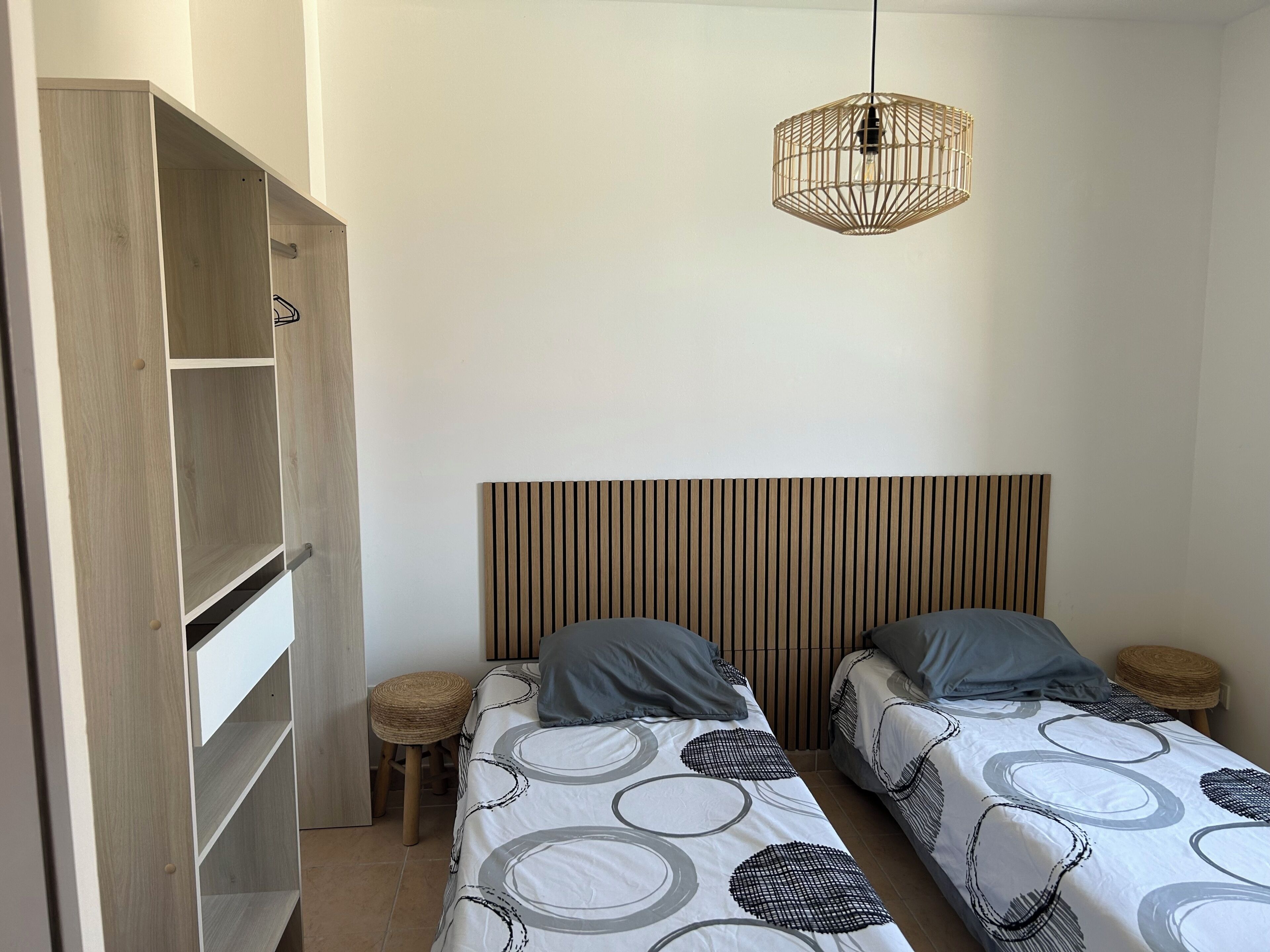 Ein Schlafzimmer mit zwei Einzelbetten, einem Holzschrank, einem gewobenen Hocker und einer Pendelleuchte.