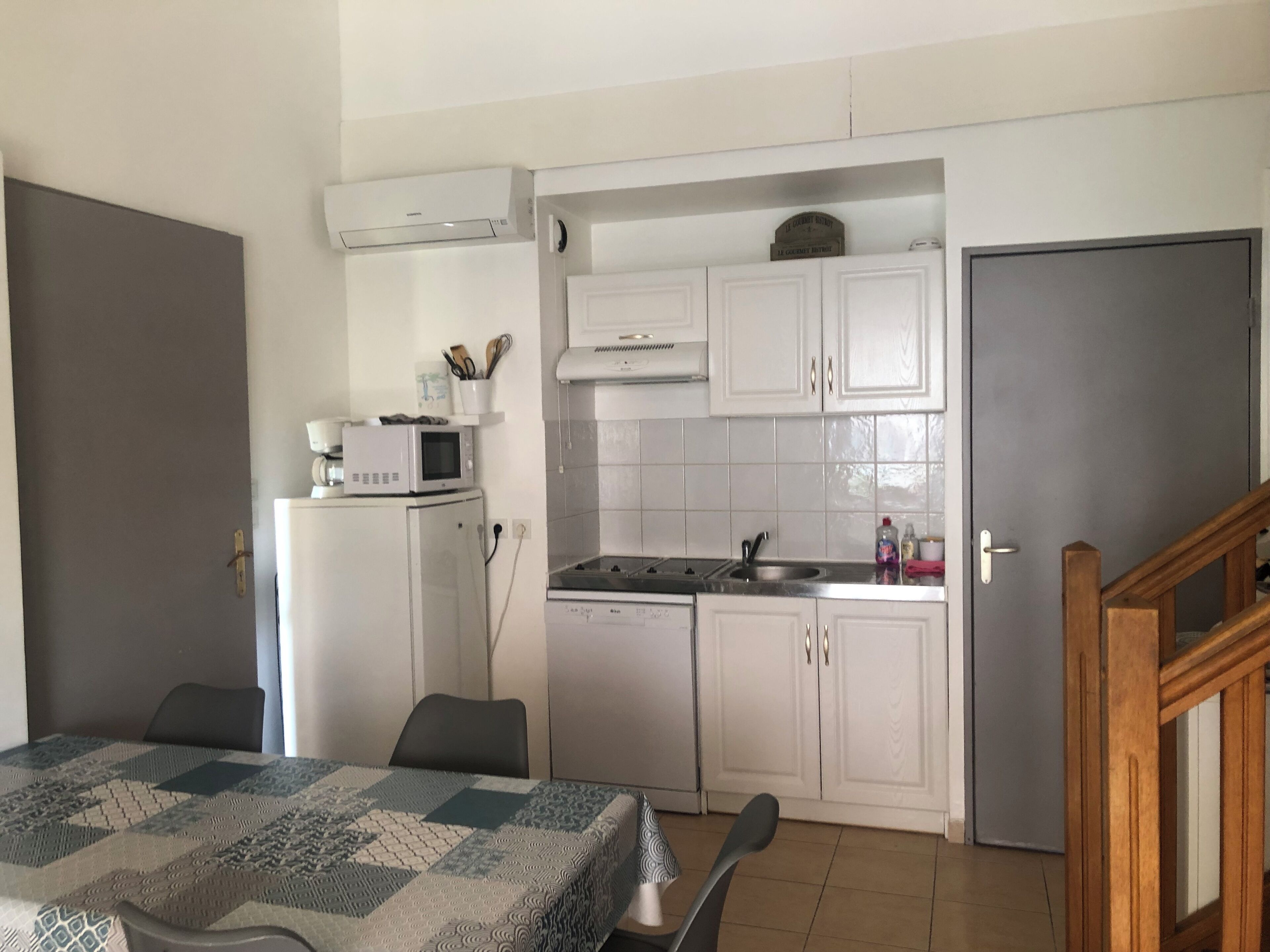 Een kleine keuken met witte kastjes, een magnetron, een spoelbak en een koelkast. Er is een eethoek met een tafel en stoelen.