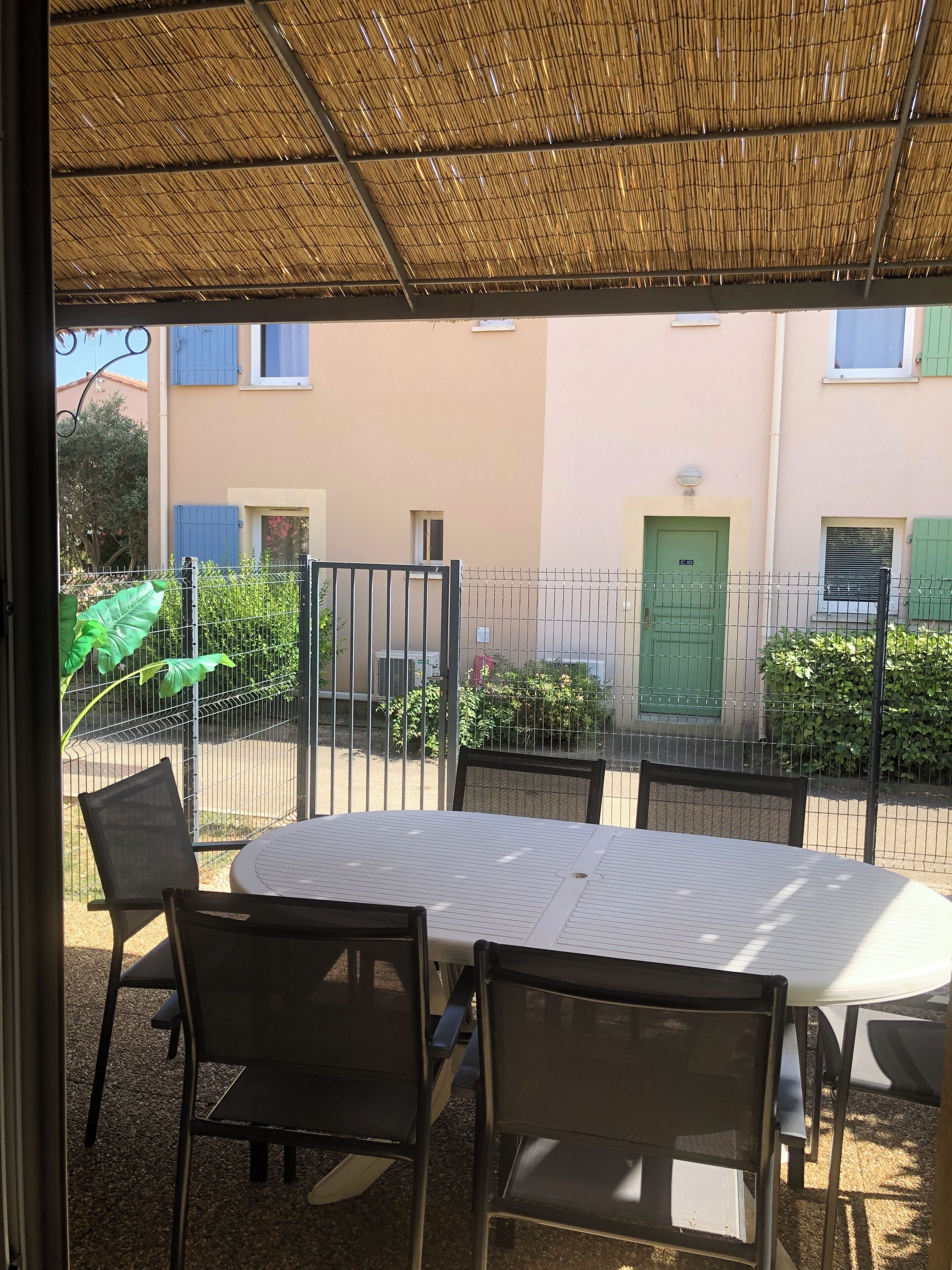 Une terrasse extérieure avec une table ronde et des chaises, un toit de chaume et une vue sur un bâtiment aux portes et fenêtres vertes.