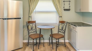 A compact kitchen with a stainless steel refrigerator, two chairs with woven seats, a small round table, and a window with blinds.