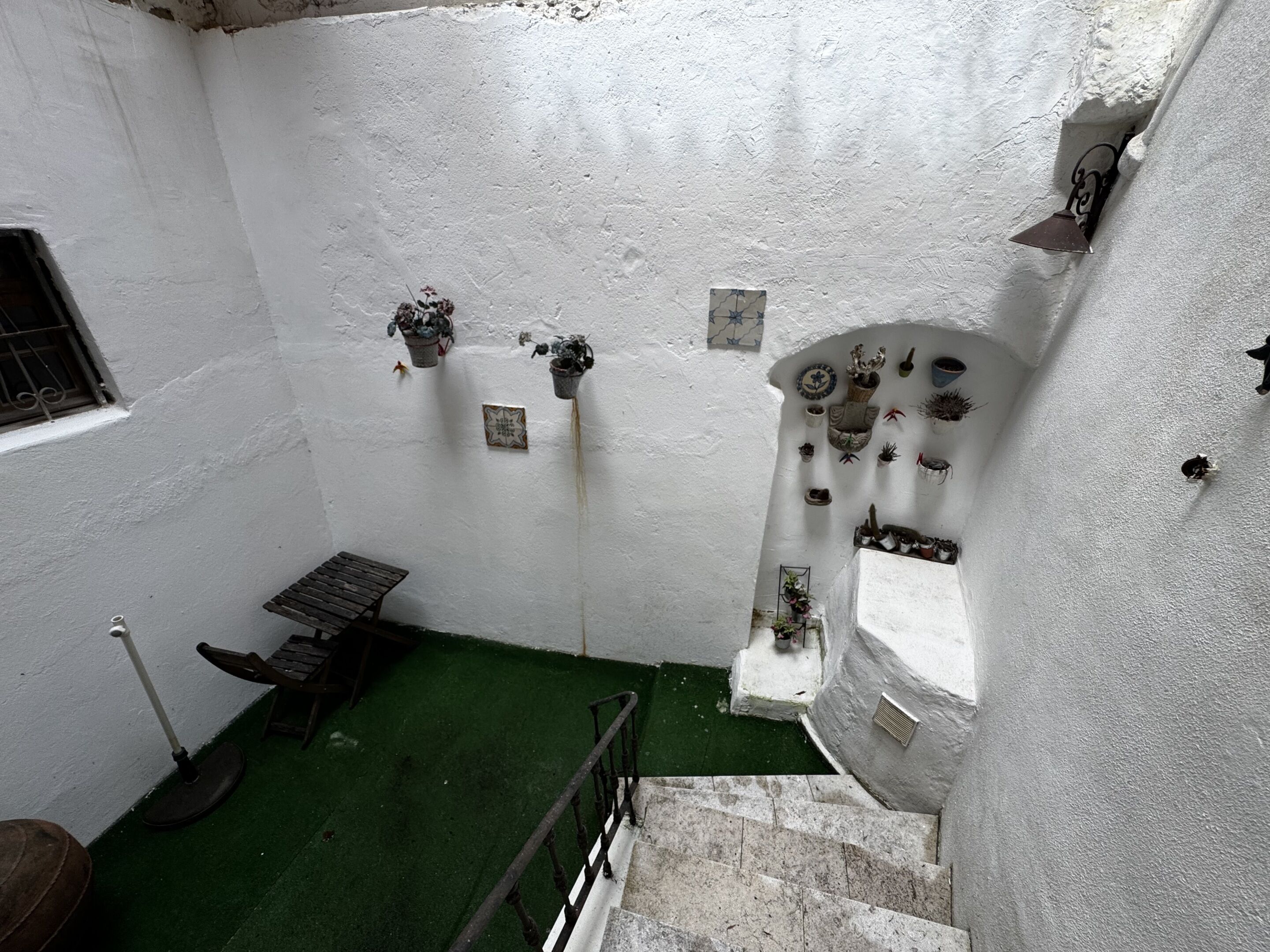 A narrow stairway leading to a room with a green artificial turf floor, white walls, and various potted plants and decorative items.