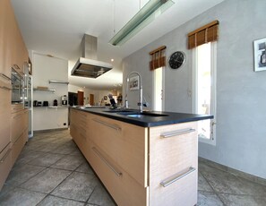 A modern kitchen with a central island, stainless steel appliances, and a large window with a view. - Villa Yoléa avec piscine privée au Grau du Roi – Vacances de rêve (Le Grau-du-Roi)