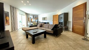 A living room with a sofa, a coffee table, and a TV. There is a kitchen in the background.