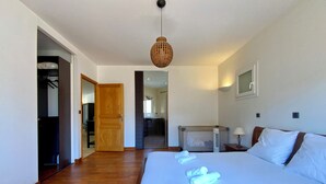 A bedroom with a wooden floor, a bed with white linens, a hanging light fixture, and a door leading to another room.