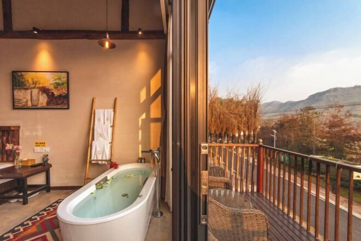 Ein modernes Badezimmer mit freistehender Badewanne, einer Holzbank, einem Gemälde und Blick auf eine gebirgige Landschaft durch große Fenster.