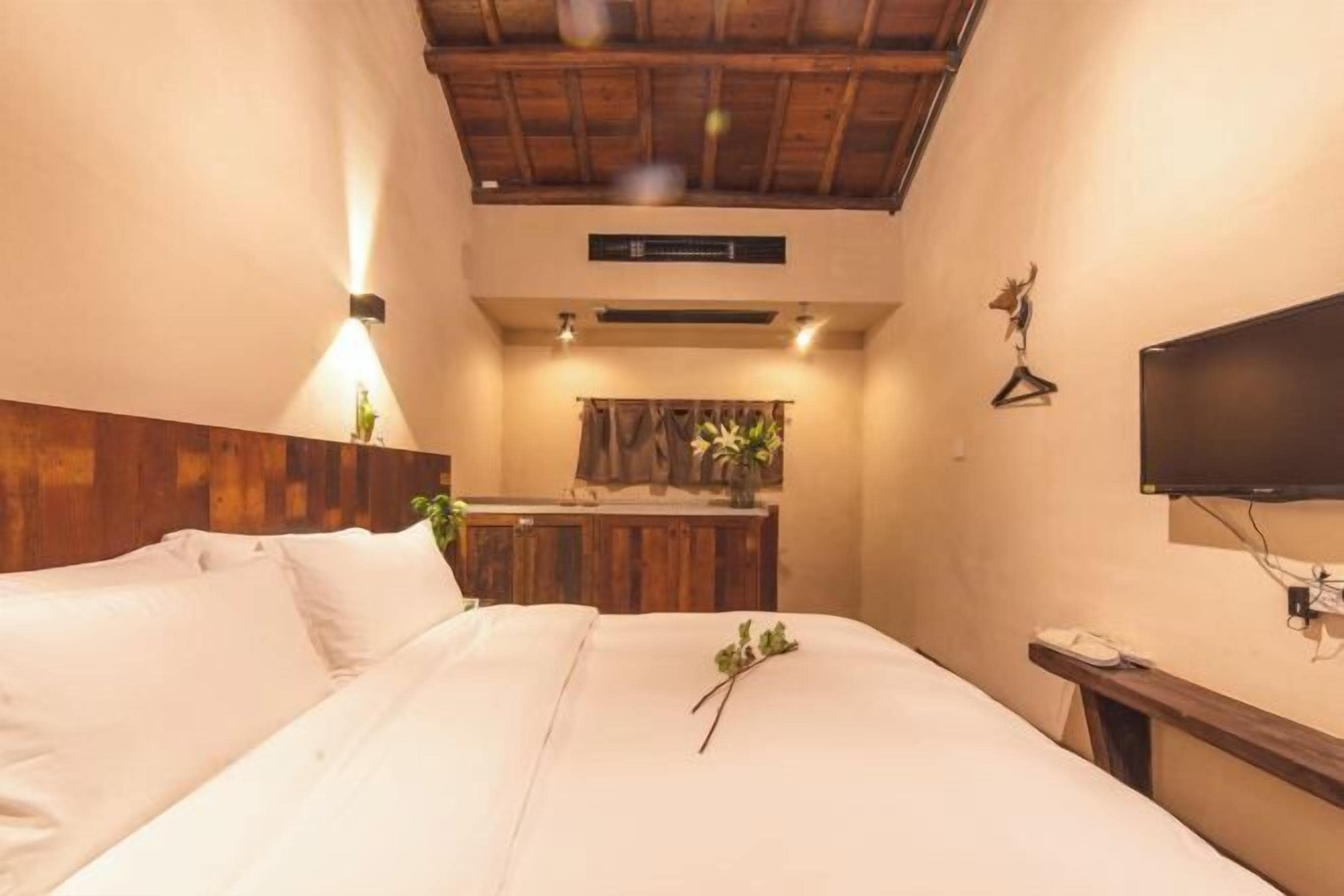 Une chambre à coucher avec une tête de lit en bois, un lit recouvert de draps blancs, une table de chevet avec une télévision et une petite plante posée sur le lit.