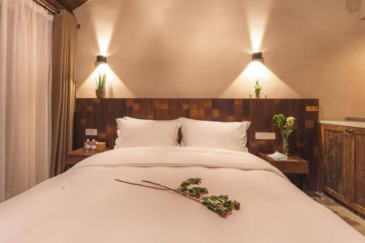 Ein Hotelzimmer mit Holz-Kopfteil, zwei Nachttischlampen, einem Bett mit weißen Leinen, einem Nachttisch mit Pflanze und einem Holzschrank.