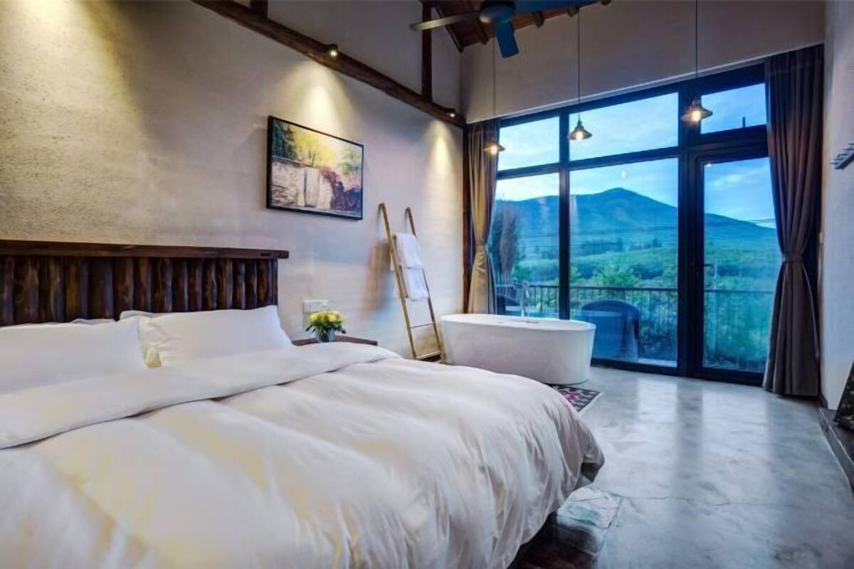 Ein Schlafzimmer mit einem großen Bett, einem Bild an der Wand, einer Leiter und Blick auf eine gebirgige Landschaft durch große Fenster.