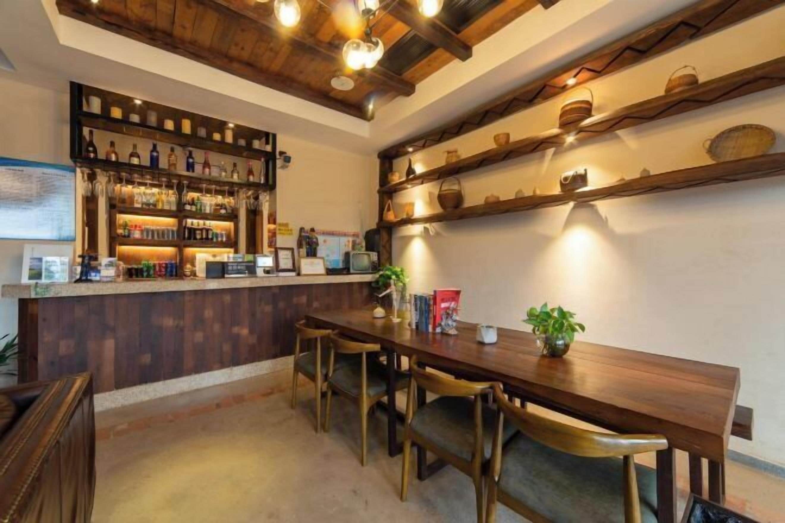 Zona de bar con estantes de madera que muestran botellas y vasos, una larga mesa de madera con sillas y una planta en maceta sobre la mesa.