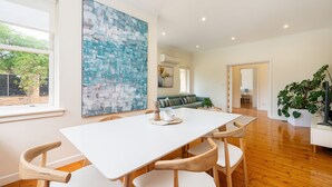 Un salon moderne avec une table à manger blanche, des chaises en bois, un grand tableau abstrait, un canapé vert et une plante en pot.