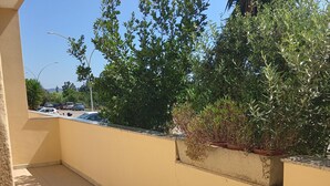 A balcony with a white table and chairs, a yellow wall, and a view of parked cars and greenery.