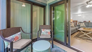 Une pièce comprenant deux fauteuils en osier avec des coussins, une petite table ronde et une porte-fenêtre coulissante donnant sur une autre pièce avec un canapé et une table basse.