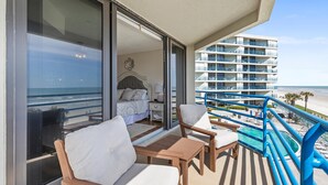 Un balcón con vista a la playa, un dormitorio con cama y mesita de noche, y una zona de piscina.