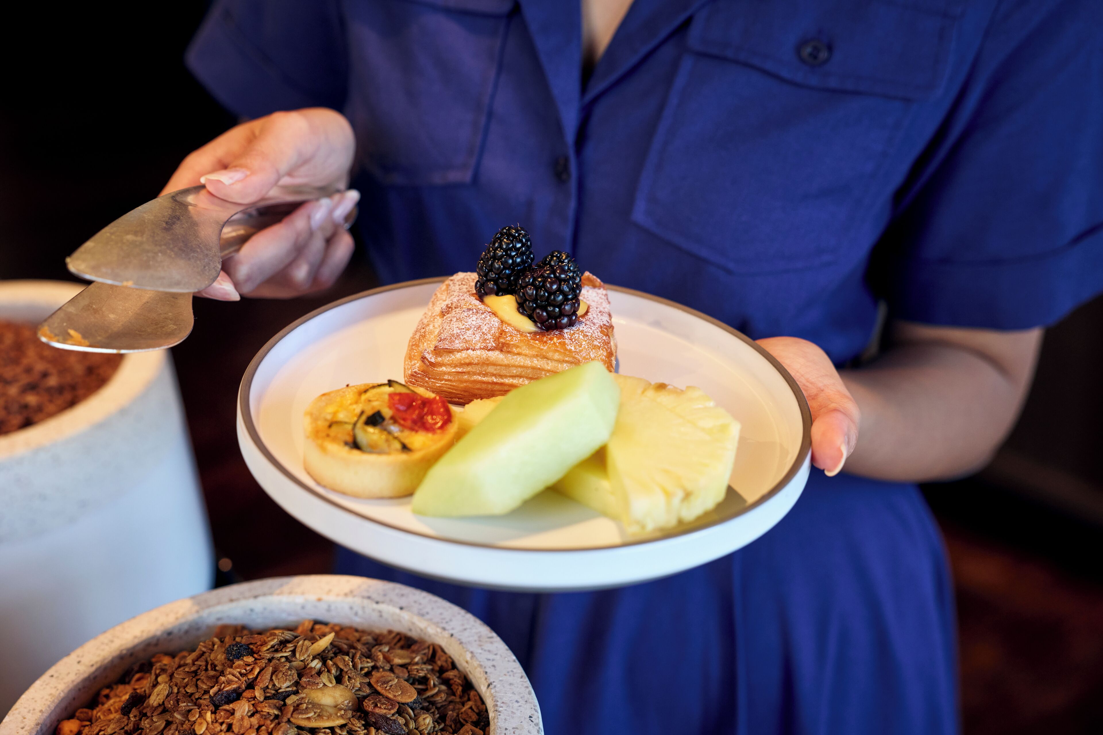 Une personne tenant une assiette sur laquelle se trouve un dessert garni de mûres, d’une tranche d’ananas et d’une cuillerée de crème.