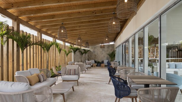A covered outdoor seating area with wicker furniture, a stone wall, and large windows. - Royal Beach Hotel Punta Cana A Jdv By Hyatt Hotel (Punta Cana)