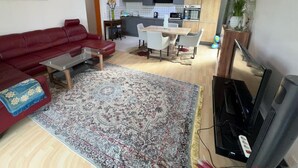 Uma sala de estar com um sofá vermelho, uma mesa de centro e uma TV. Há uma área de jantar com mesa e cadeiras, e uma cozinha com eletrodomésticos modernos.